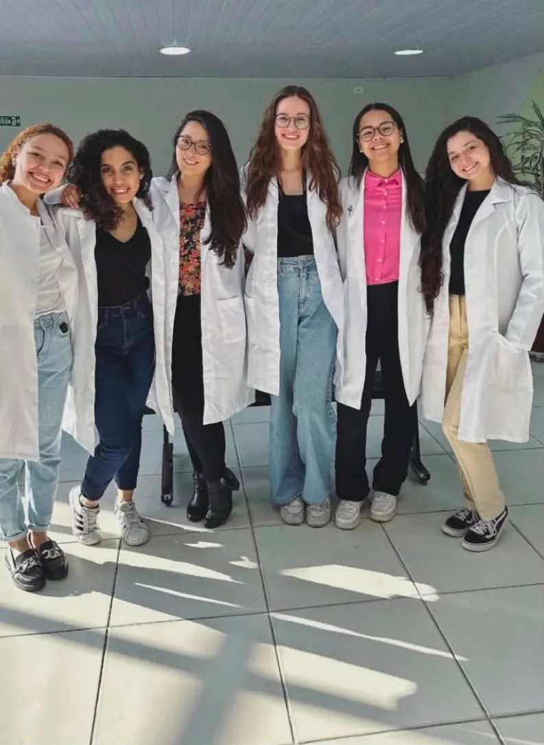 Seis integrantes da equipe de jaleco branco posam abraçadas e sorrindo.