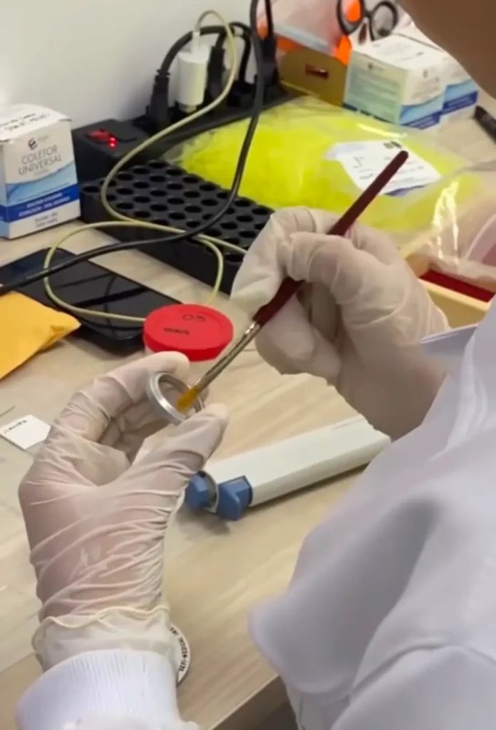 Mãos com luvas manipulam material em bancada de laboratório.